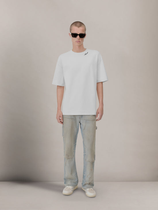 White crew neck t-shirt with script lettering on shoulder, worn with light blue jeans.