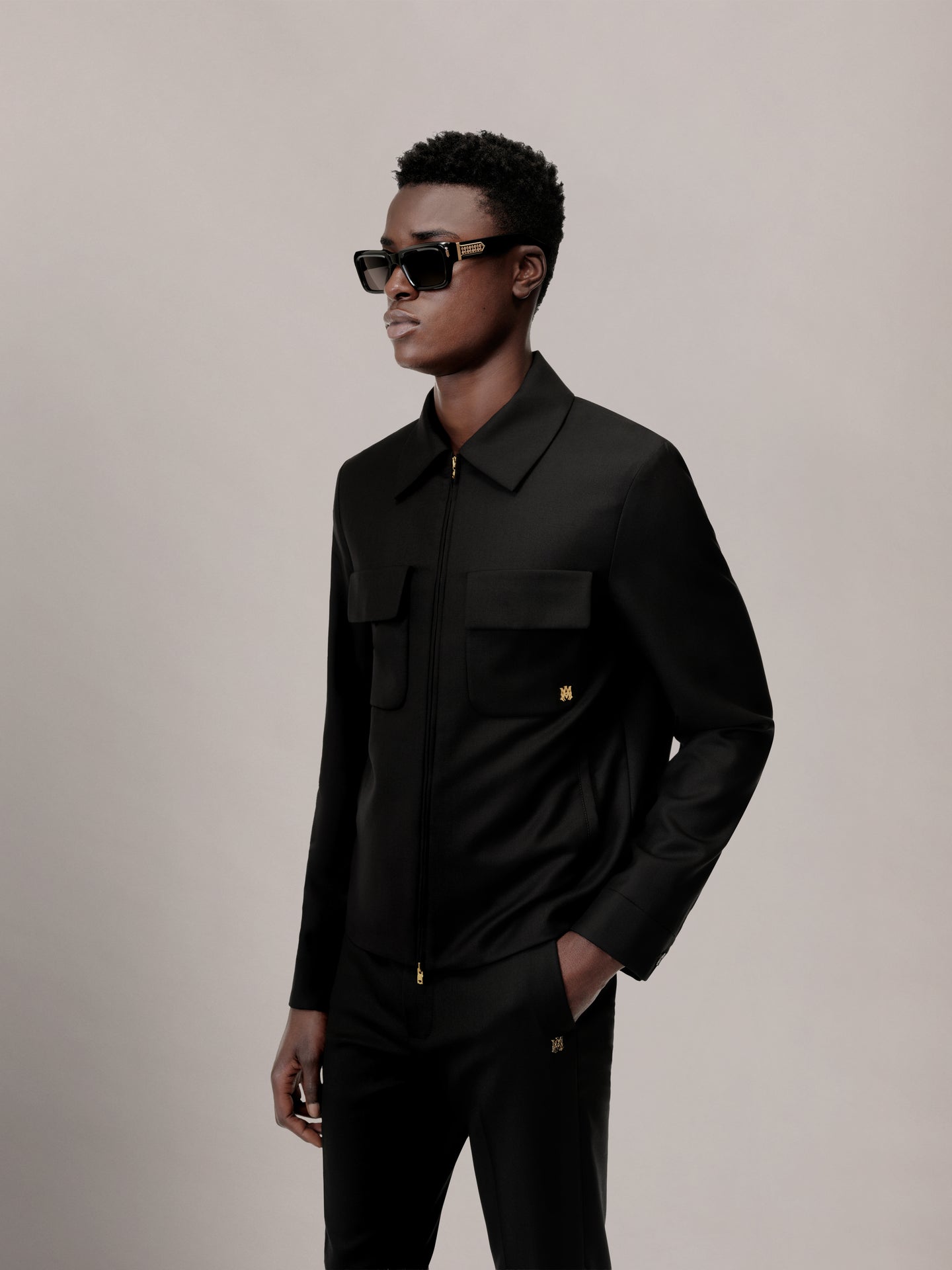 Black blouson jacket with collar, zip front, chest pockets, and gold hardware worn by model.