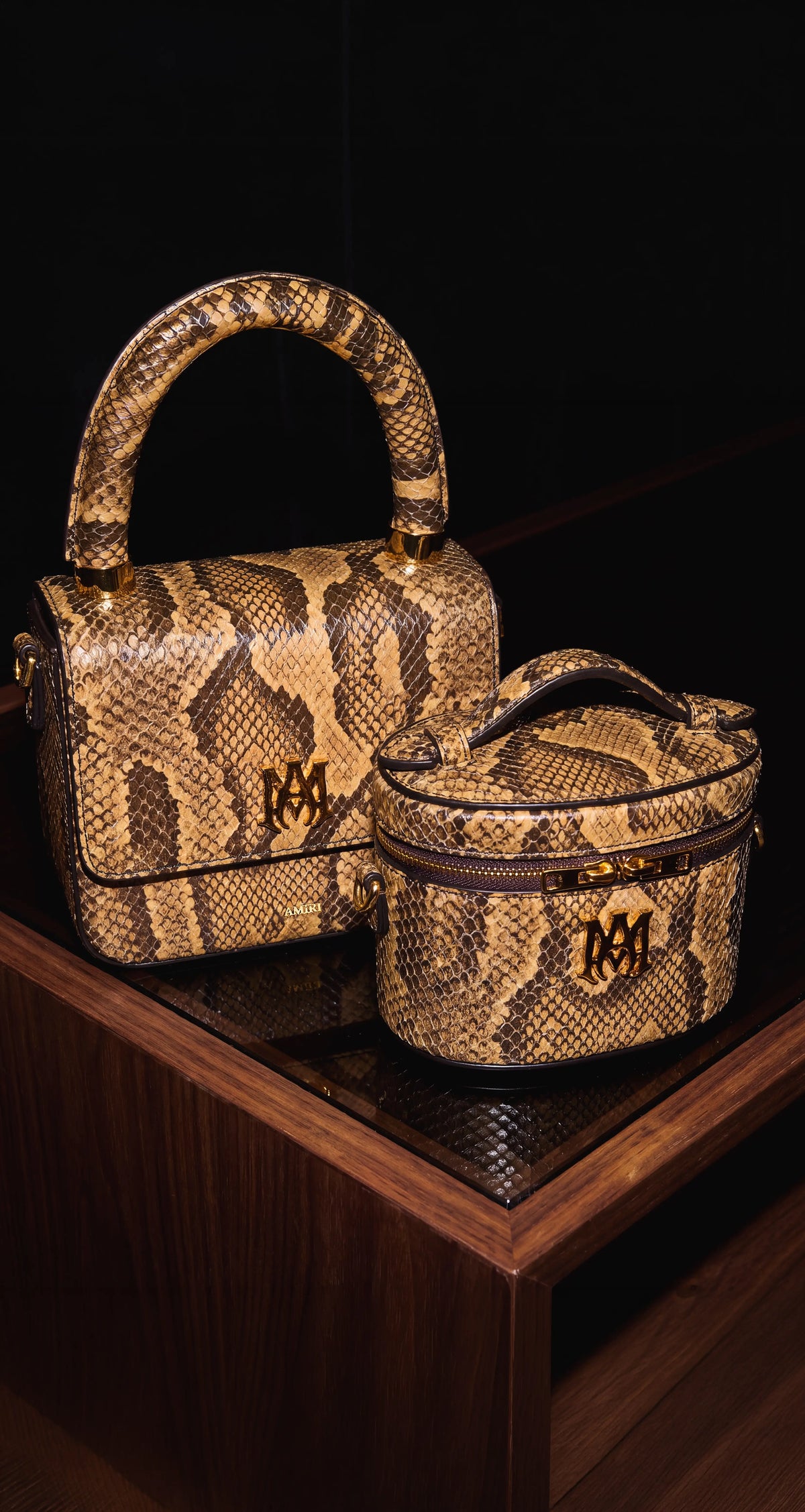 Two snakeskin-patterned handbags displayed on a dark wooden surface against a black background. The larger bag is structured and rectangular with top handles, and the smaller bag is a rounded cylindrical case with a top handle and zipper closure.